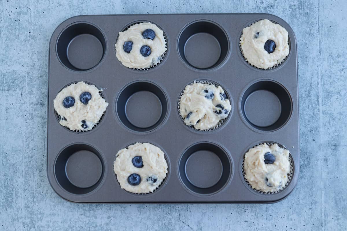 Muffin tray with five cups filled with blueberry muffin batter, placed in a checkerboard pattern. The tray is on a textured gray surface.