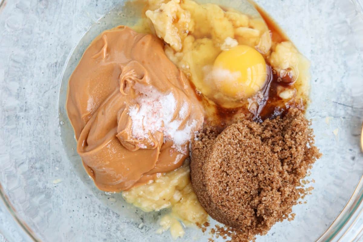 A glass bowl containing peanut butter, mashed banana, an egg, brown sugar, salt, and vanilla extract. The ingredients are partially mixed, ready for baking preparation.