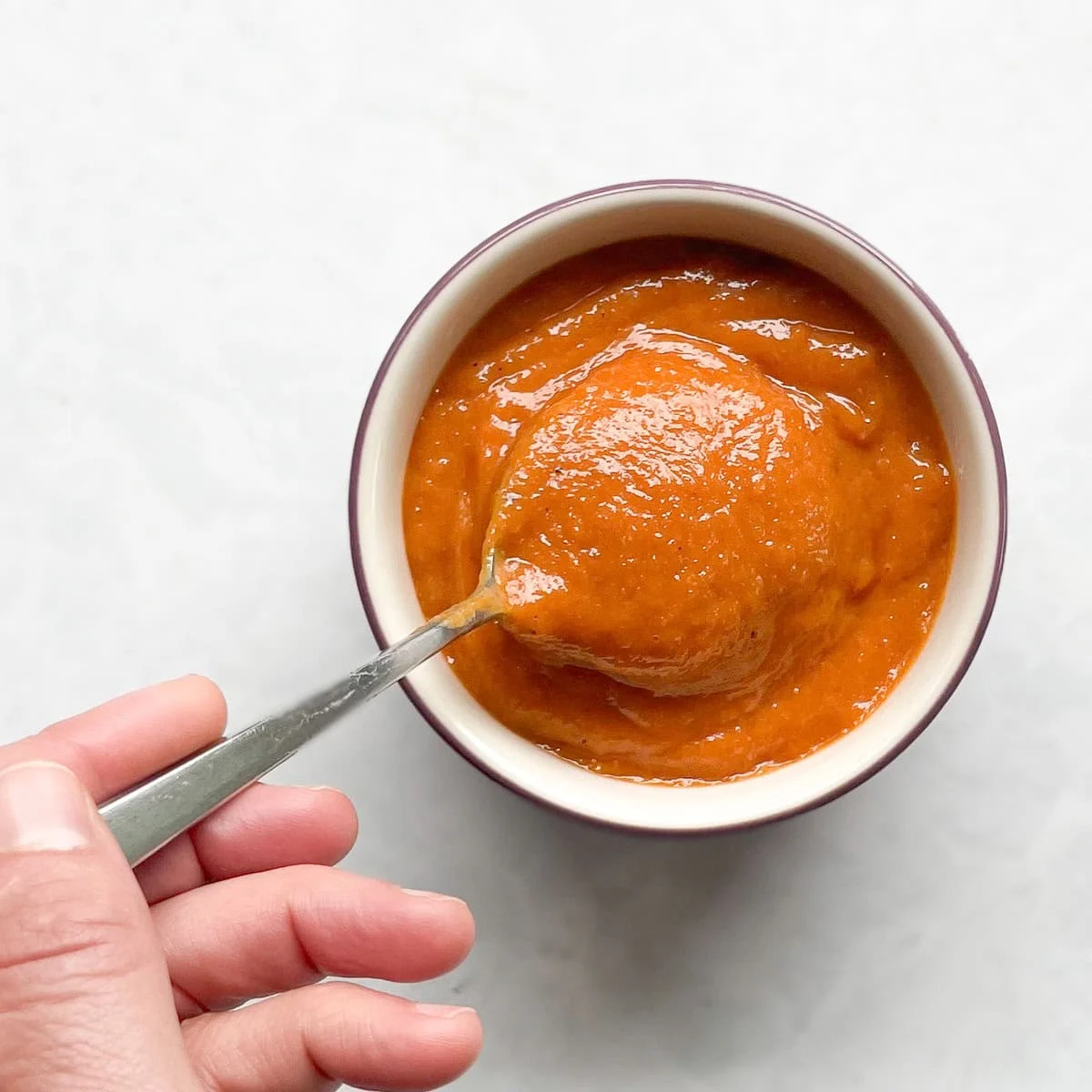 A hand holding a spoon is shown dipping into a small bowl filled with thick, smooth red sauce. The bowl is placed on a light-colored surface, and the sauce appears to have a rich, glossy texture.