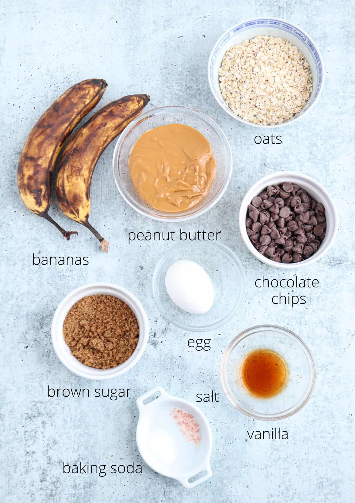 Overhead view of baking ingredients on a light surface: bananas, peanut butter, oats, chocolate chips, brown sugar, an egg, salt, vanilla extract, and baking soda. Each item is labeled.