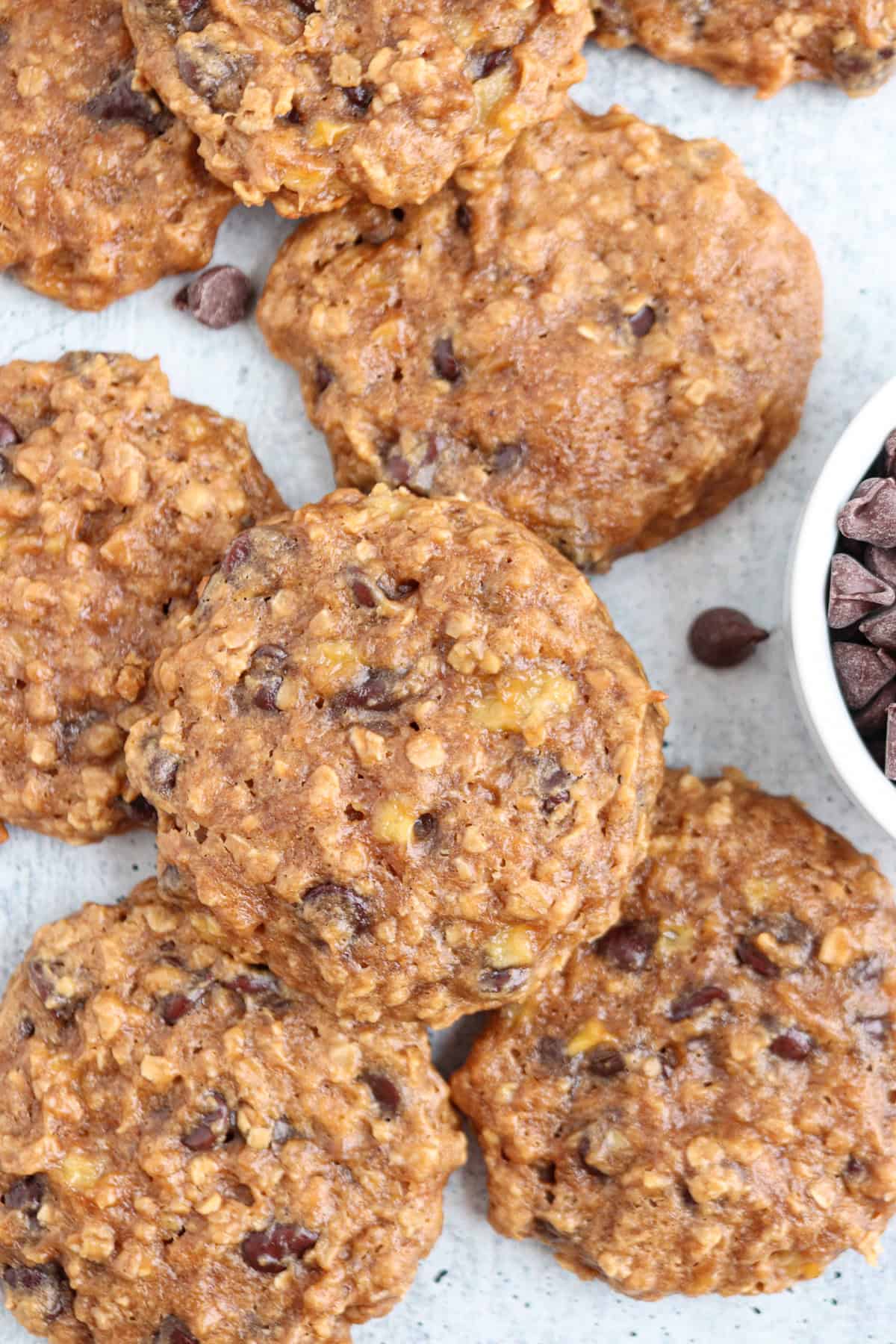 A pile of oatmeal chocolate chip cookies is displayed on a light surface. The cookies are golden brown with visible oats and chocolate chips scattered throughout. A small white bowl filled with chocolate chips is partially visible on the right side.