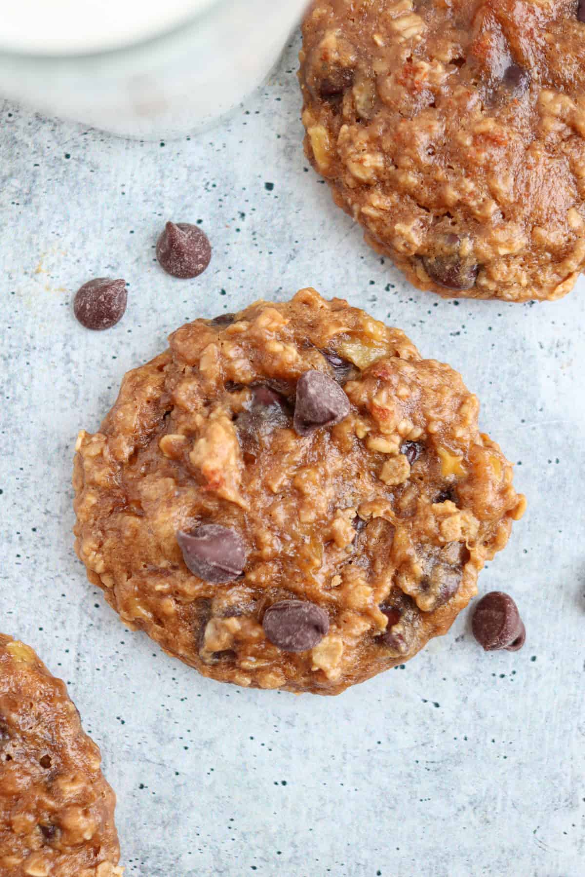 A close-up of a chocolate chip oatmeal cookie on a gray surface. The cookie has visible oats, chocolate chips, and a rustic texture. Nearby are a few scattered chocolate chips and partial glimpses of other cookies and a glass of milk.