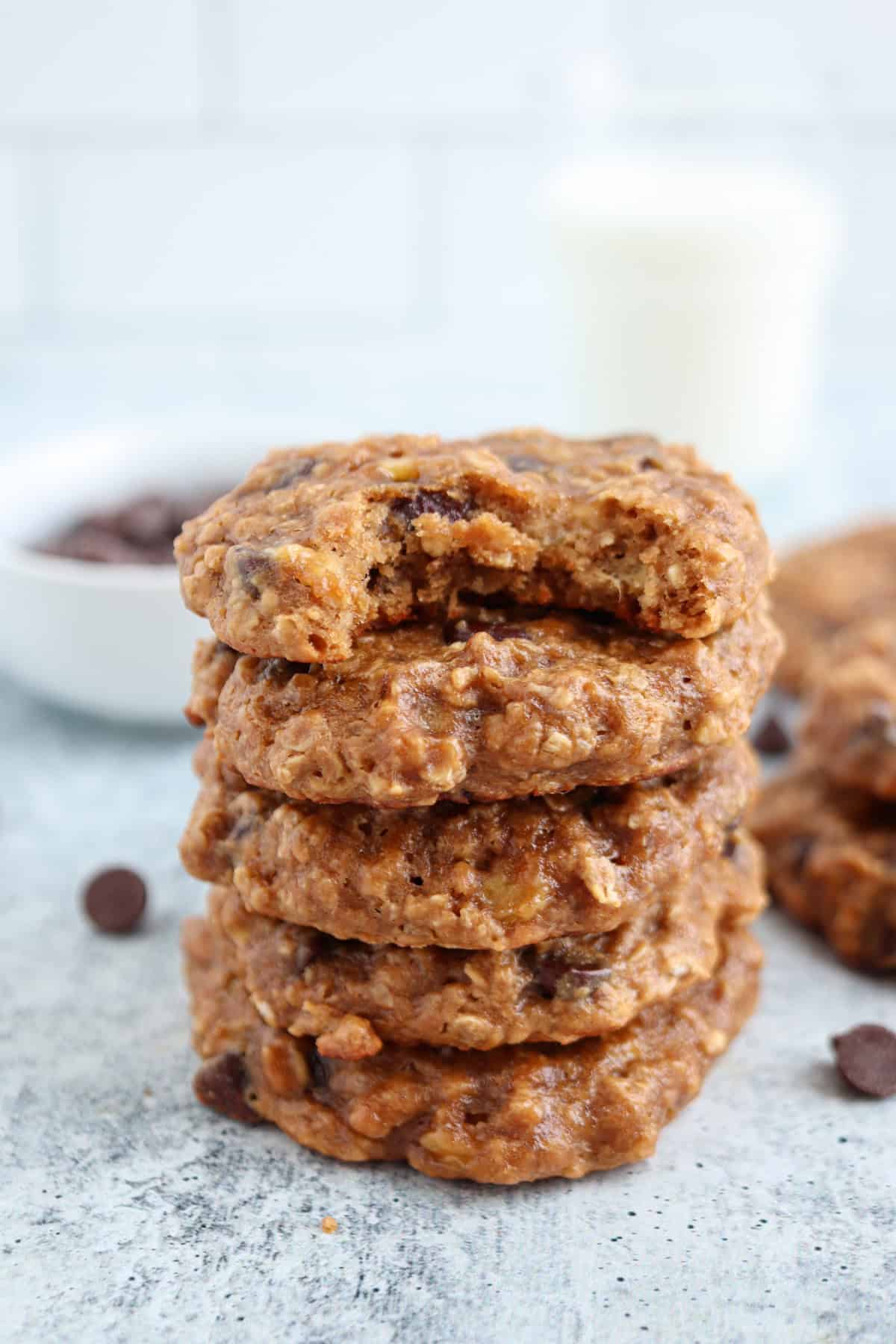 Stack of five oatmeal chocolate chip cookies with a bite taken from the top one, on a light textured surface. Background includes a bowl of chocolate chips and a glass of milk.