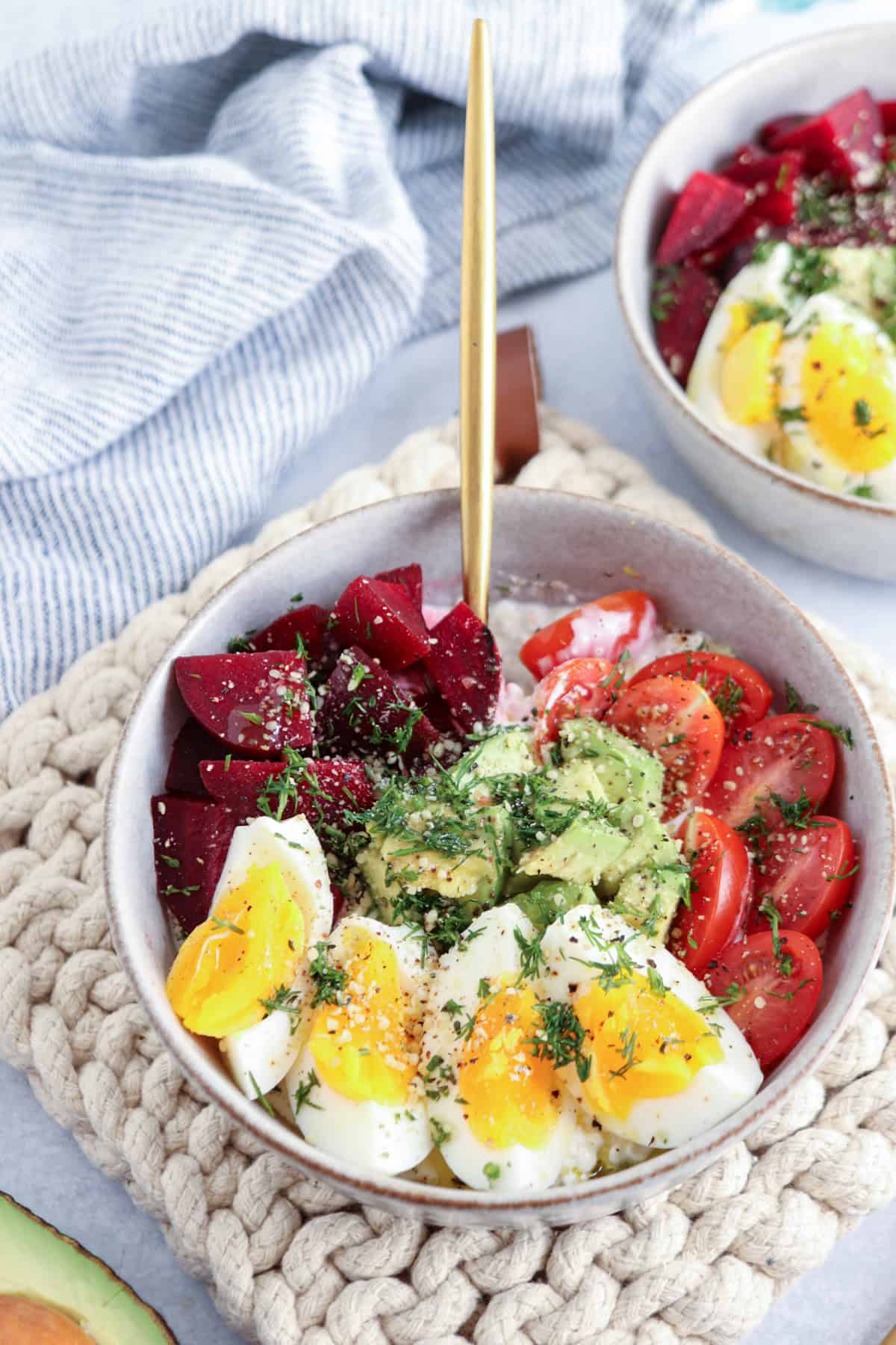 A colorful bowl of sliced boiled eggs, cherry tomatoes, beetroot, and mashed avocado garnished with herbs. The bowl is placed on a knitted placemat with a fork. A partial view of another similar bowl is in the background.
