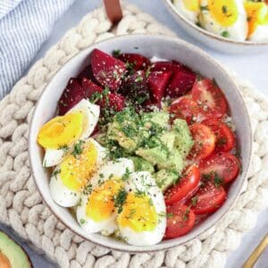A bowl with sliced boiled eggs, cherry tomatoes, avocado, and beetroot, sprinkled with herbs and seeds. The bowl is on a textured, knitted mat, with another similar dish partially visible in the background.