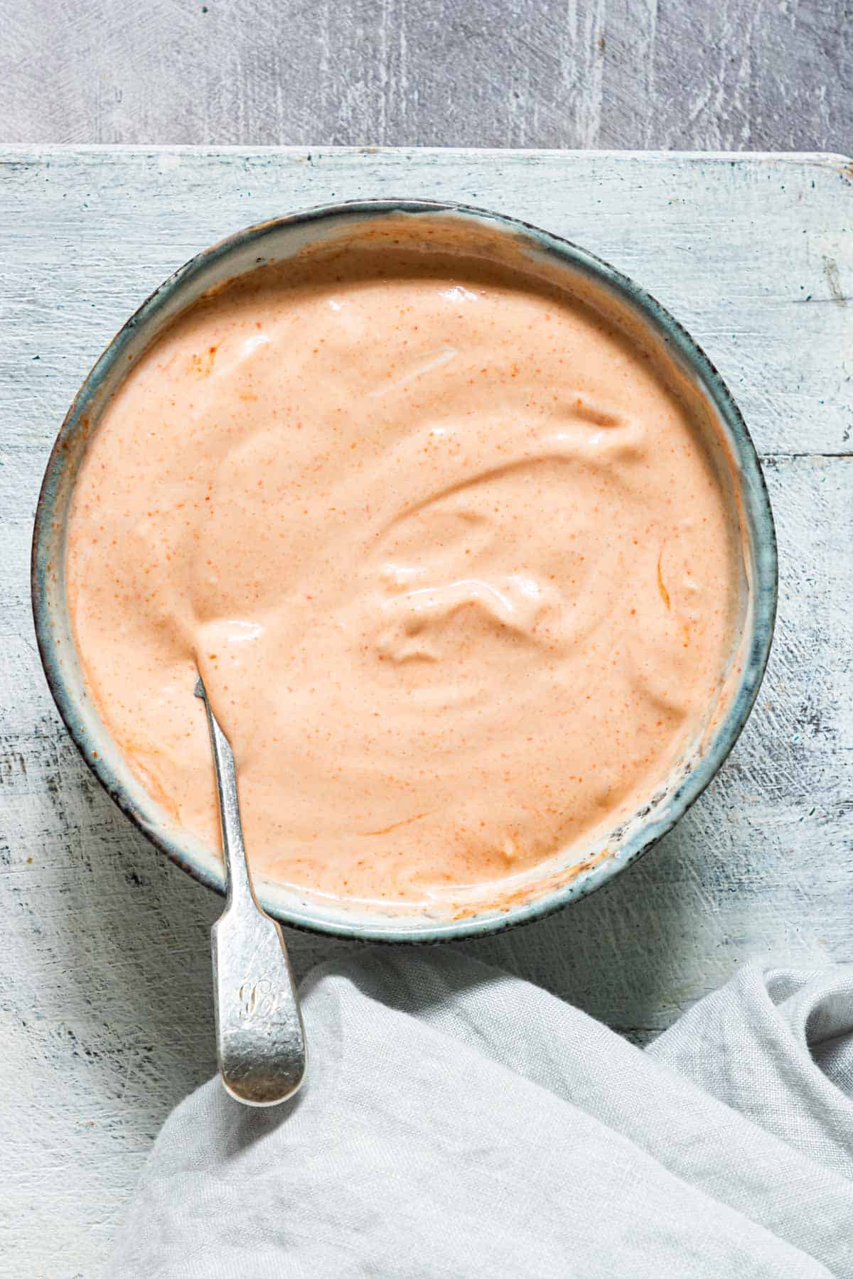 A bowl of creamy, orange dipping sauce sits on a light wooden surface. A spoon is resting in the bowl, with a cloth nearby. The sauce appears smooth and slightly speckled.