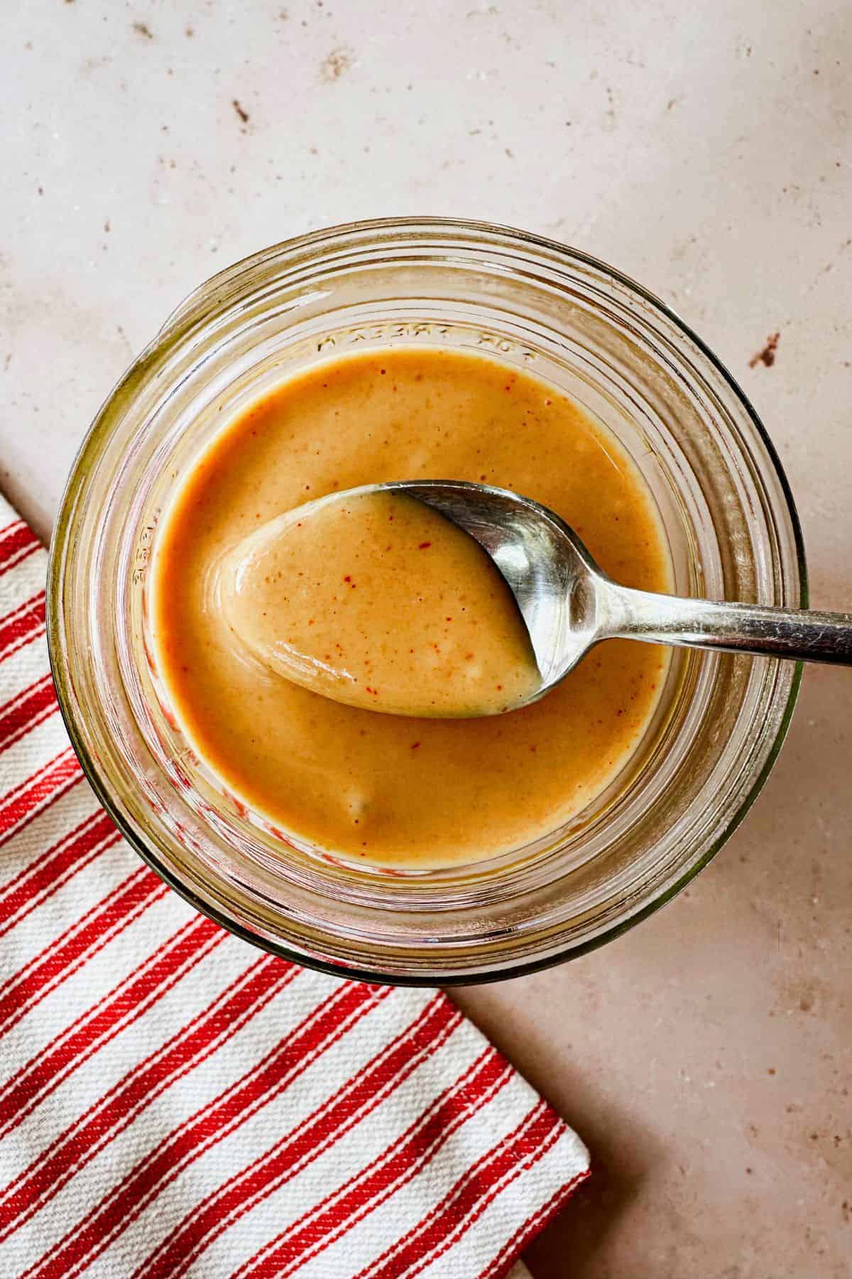 A glass jar filled with creamy peanut sauce, with a spoon resting inside. The jar is placed on a light surface next to a red and white striped cloth.