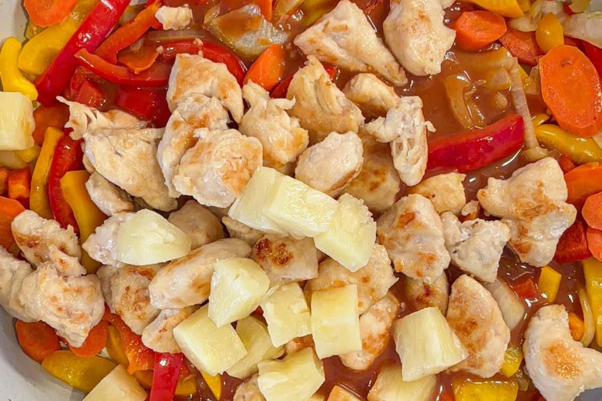 A close-up of a colorful stir-fry featuring chunks of chicken, pineapple pieces, and a mix of vegetables including red and yellow bell peppers, and carrots, all coated in a glossy sauce.