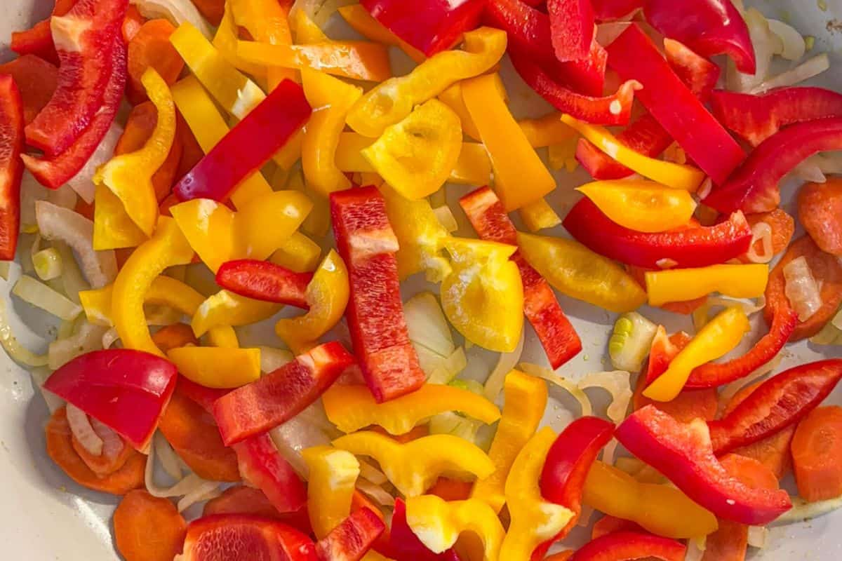 A close-up of sliced red and yellow bell peppers mixed with carrot slices on a white surface, illuminated by warm light.