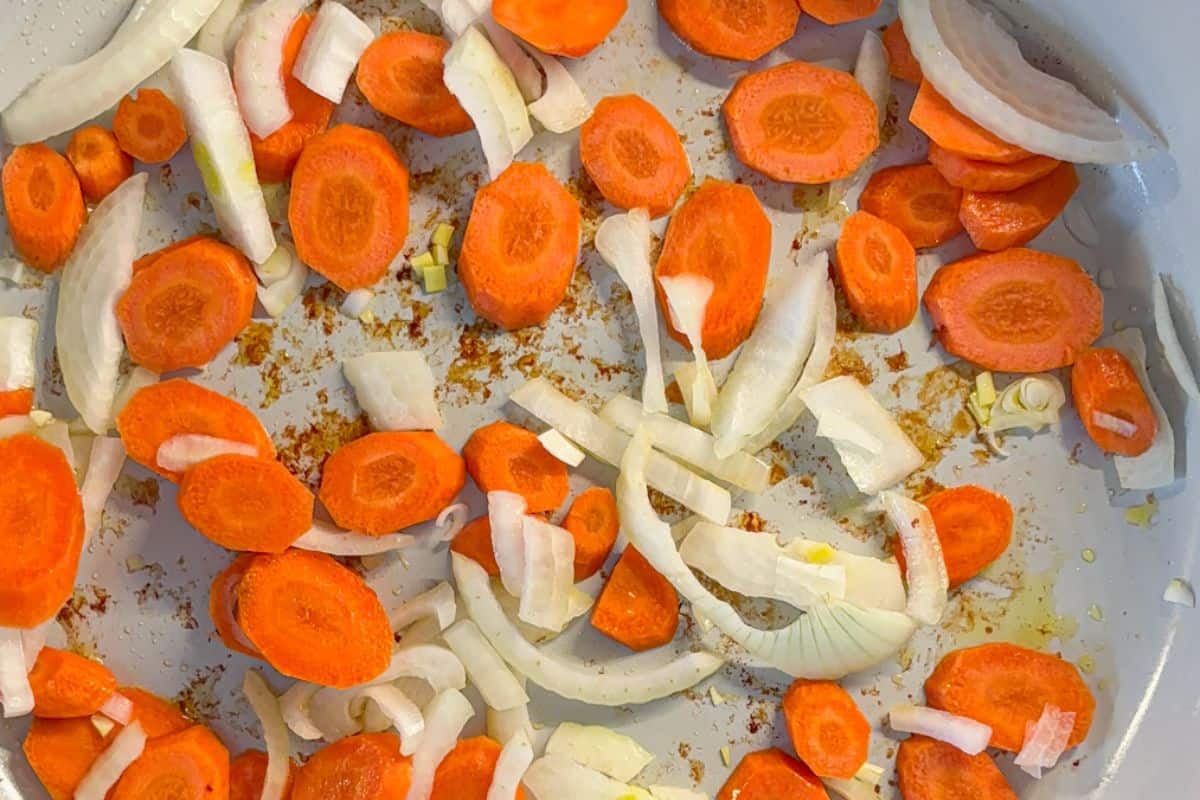 Sliced carrots and onions sautéing in a pan with a touch of oil, showing browning on the vegetables. The pan has a light coating of spices and oil, creating a colorful and appetizing scene.