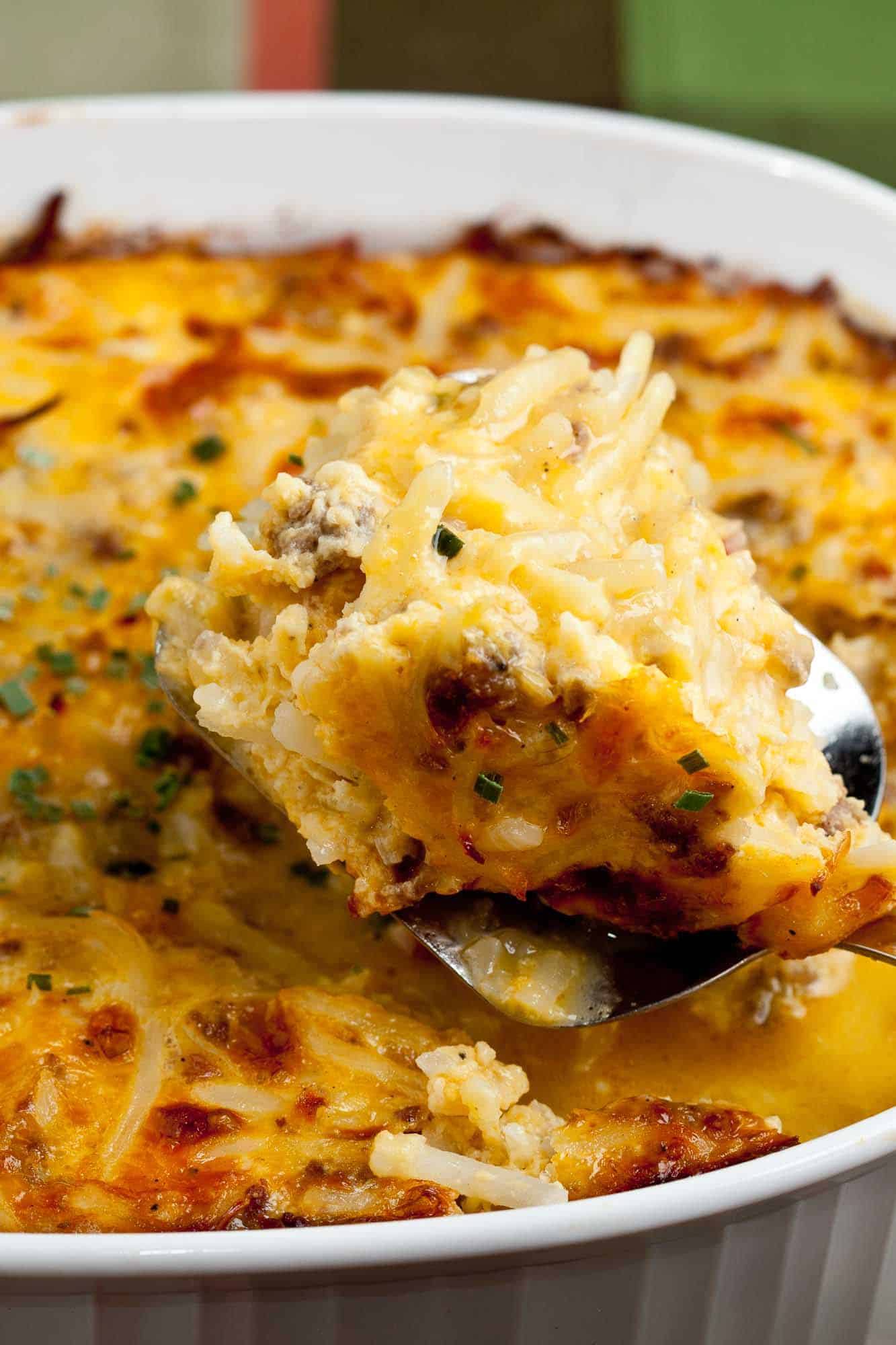 A close-up of a cheesy hash brown casserole in a white dish. A spoon is lifting out a portion, showing melted cheese, ground meat, and chives. The top is golden and crispy.