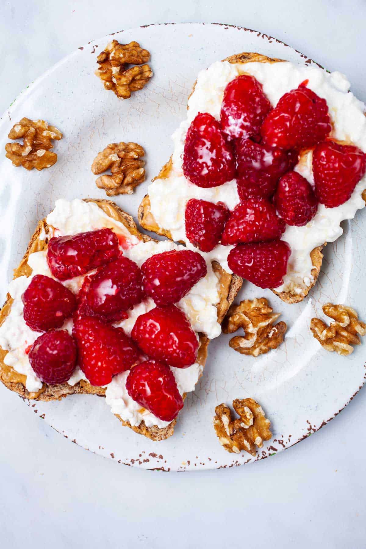 Two slices of toast topped with cottage cheese and fresh raspberries are on a plate. Walnuts are scattered around the toast, adding a rustic touch to the presentation.