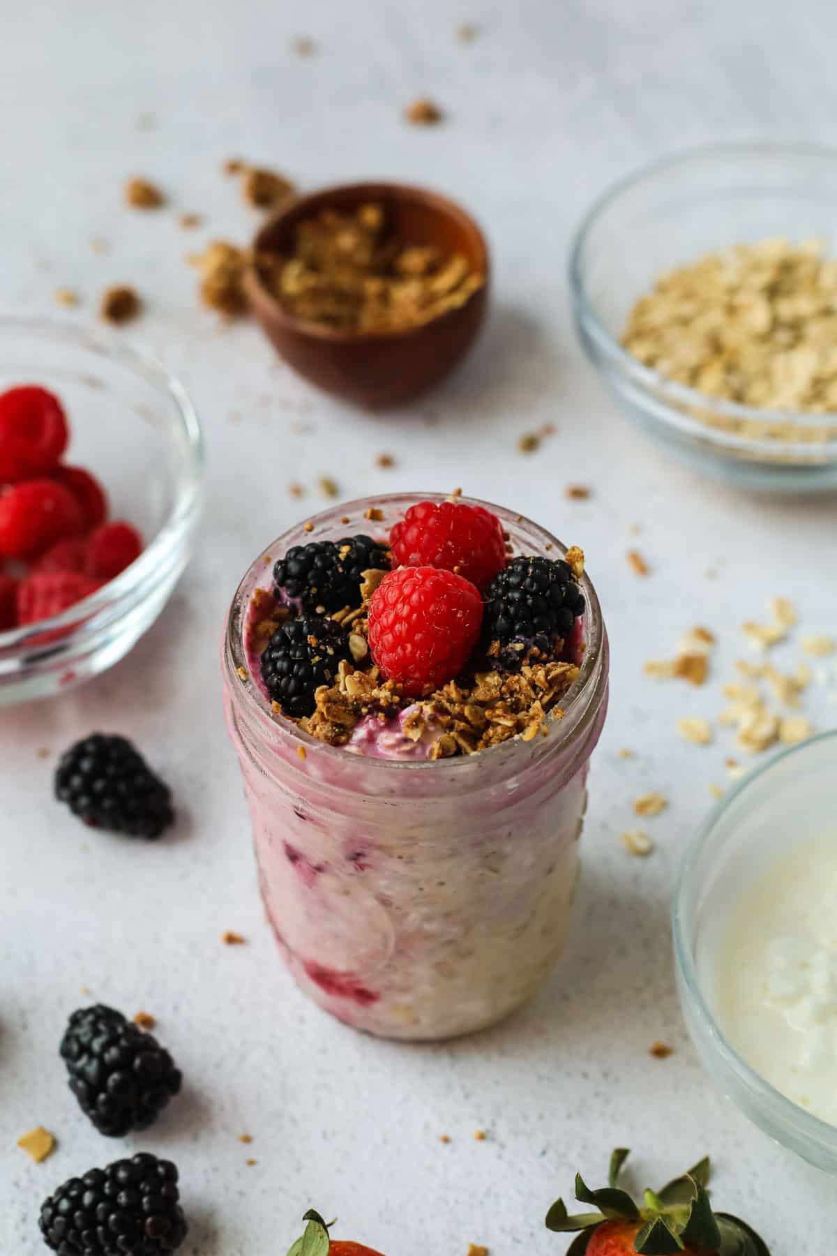 A mason jar of yogurt topped with granola, raspberries, and blackberries is surrounded by bowls of oats, granola, raspberries, and yogurt on a light textured surface. Sprinkled oats and berries are scattered around.