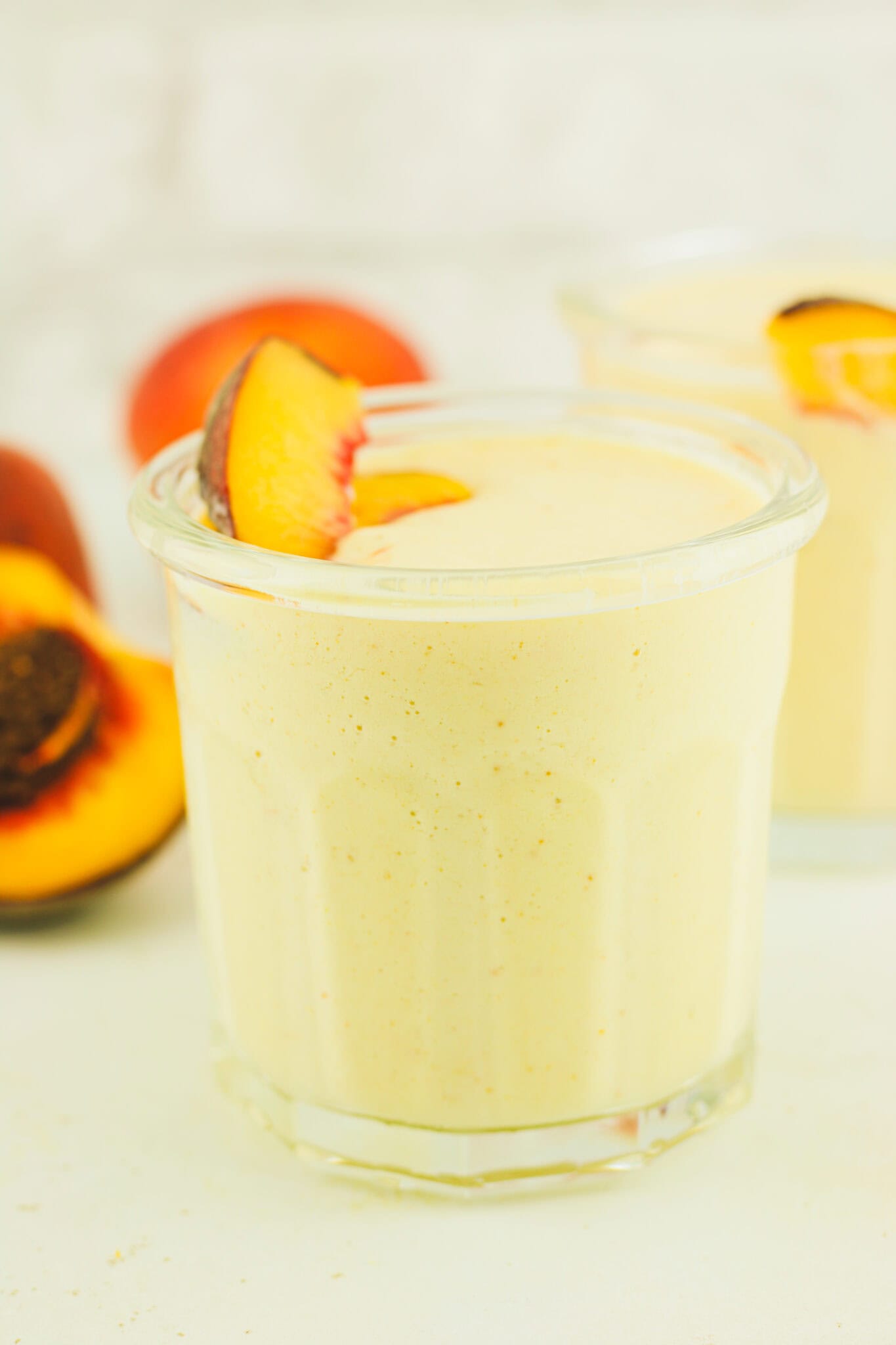 A creamy peach smoothie in a clear glass, garnished with a slice of fresh peach on the rim. In the blurred background, whole peaches are visible on a light surface.