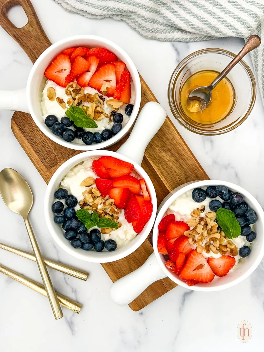 Three bowls of yogurt topped with strawberries, blueberries, walnuts, and mint leaves are arranged on a wooden board. A small glass bowl filled with honey and a honey dipper sits nearby. Two gold spoons are placed alongside.