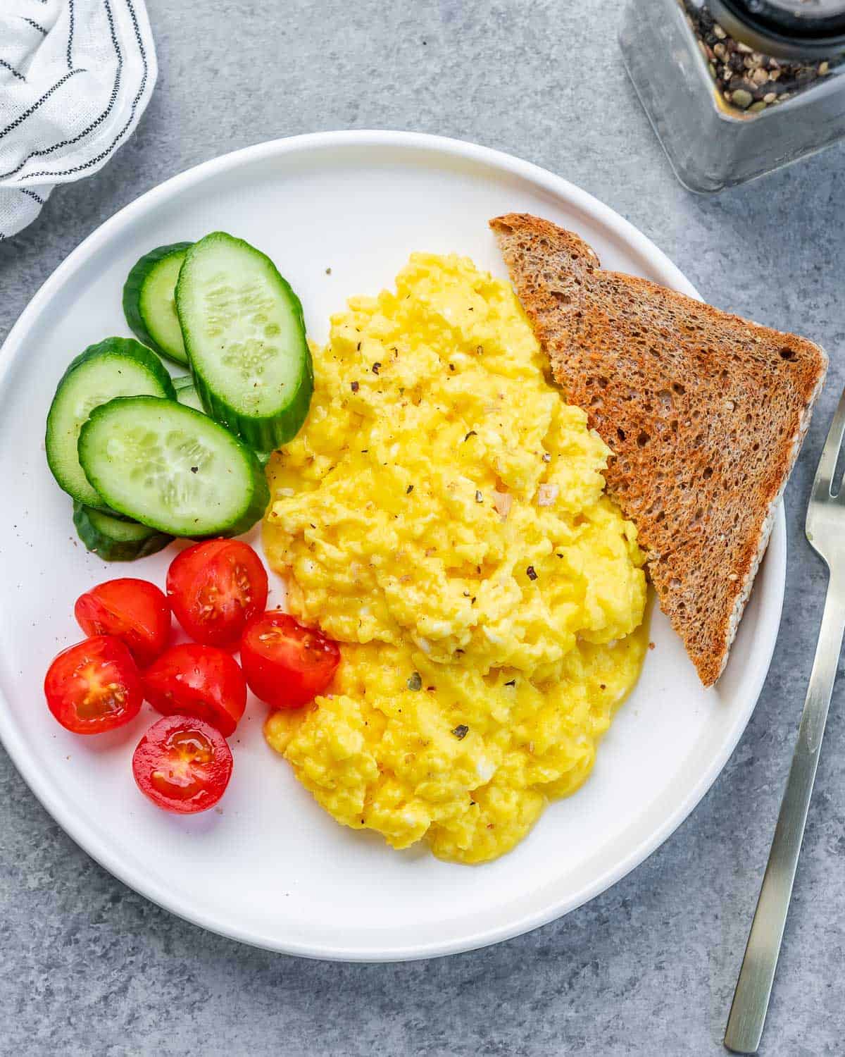 A white plate with scrambled eggs, a slice of brown toast, sliced cucumbers, and halved cherry tomatoes. A fork is placed beside the plate on a gray surface. A pepper shaker and part of a striped napkin are visible nearby.