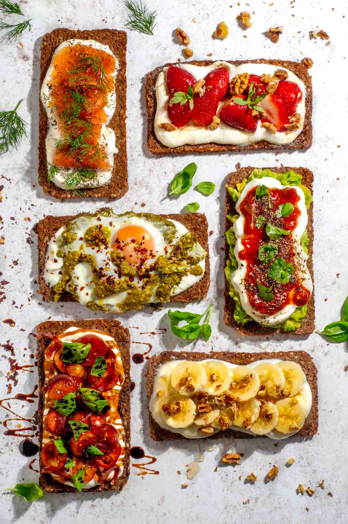 Six slices of toast with a variety of toppings: smoked salmon with dill, strawberries with cream, avocado and egg, tomato and mozzarella, kiwi and balsamic, and banana with walnuts, arranged on a white surface with scattered herbs.