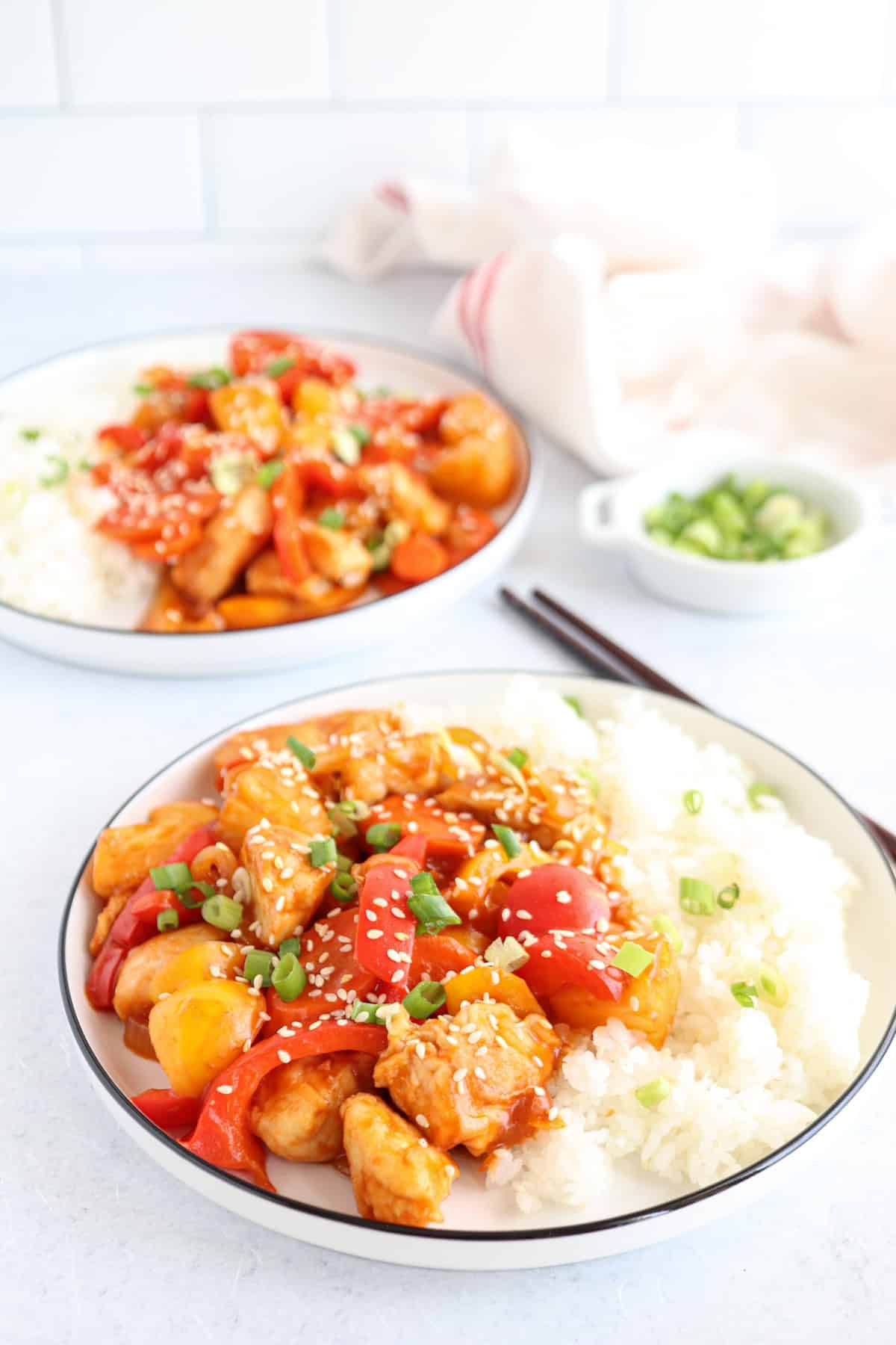 Two plates of sweet and sour chicken with bell peppers and sesame seeds, served alongside white rice. Garnished with chopped green onions. A small bowl with additional green onions is in the background. Chopsticks rest near the plates.