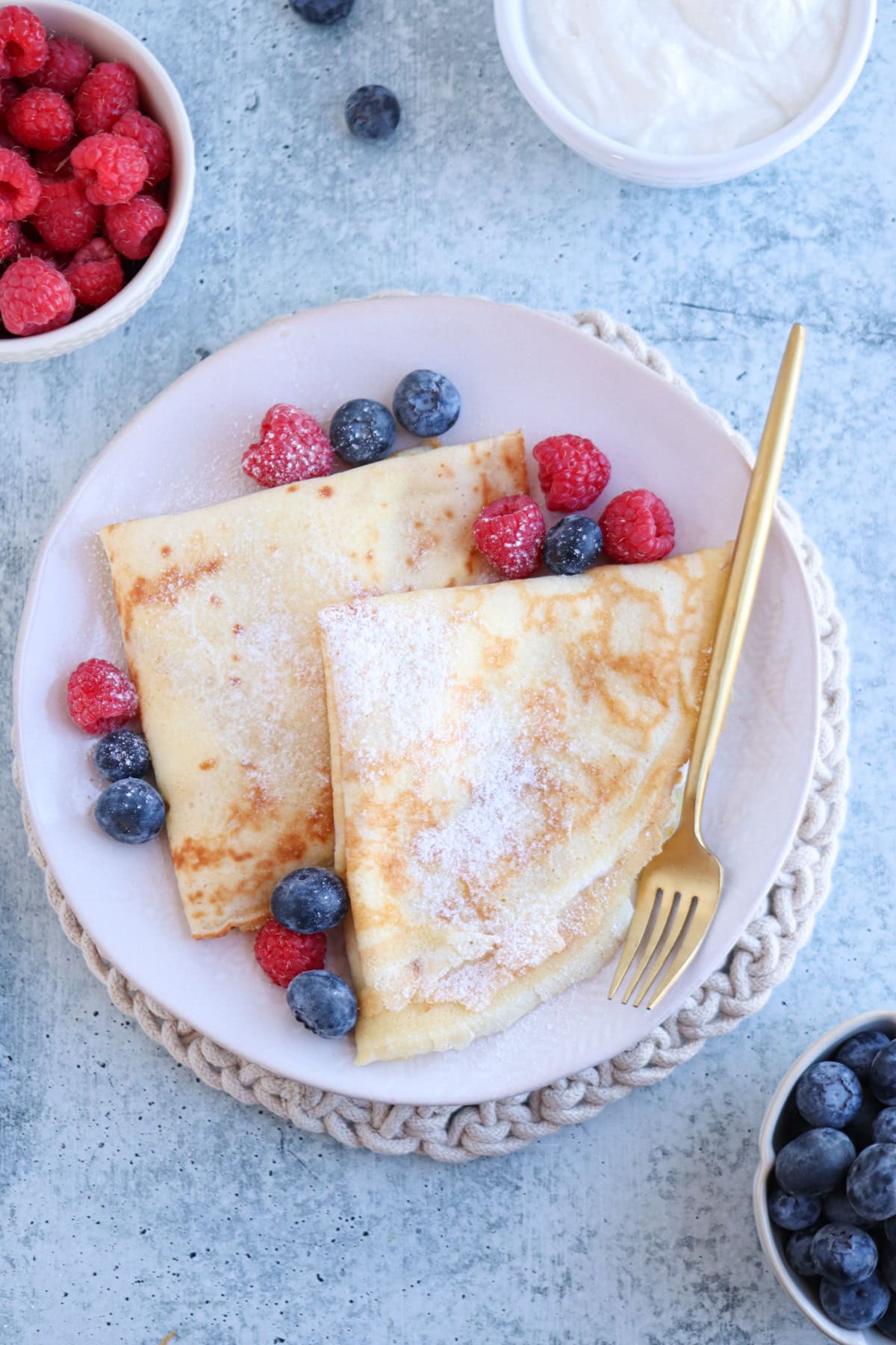 A plate of crepes, reminiscent of a classic nalesniki recipe, is folded and sprinkled with powdered sugar, garnished with raspberries and blueberries. A gold fork rests on the plate, while additional berries and a bowl of cream adorn the gray surface nearby.