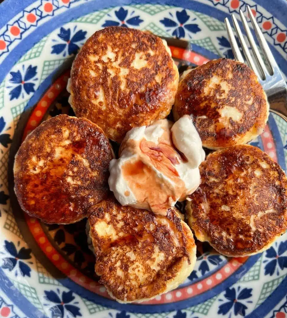 A plate with five golden-brown pancakes arranged around a dollop of cream with a drizzle of sauce. The plate has a decorative blue and red pattern, and a fork is placed on the side.