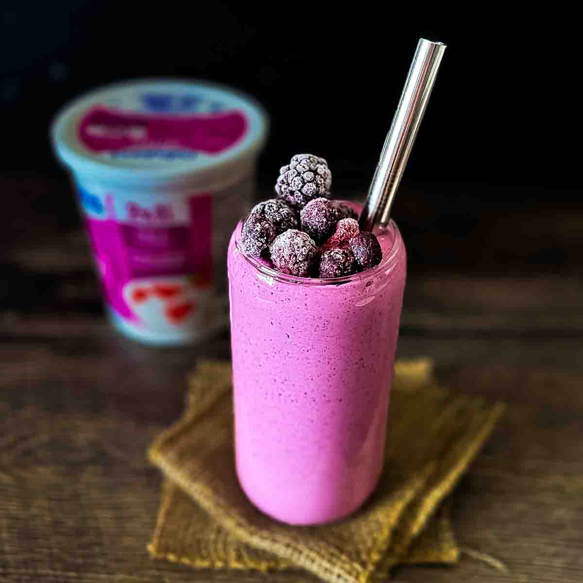 A pink smoothie topped with frozen blackberries in a glass with a metal straw, placed on a piece of burlap. In the blurred background, there is a container of yogurt. The surface is dark wood.