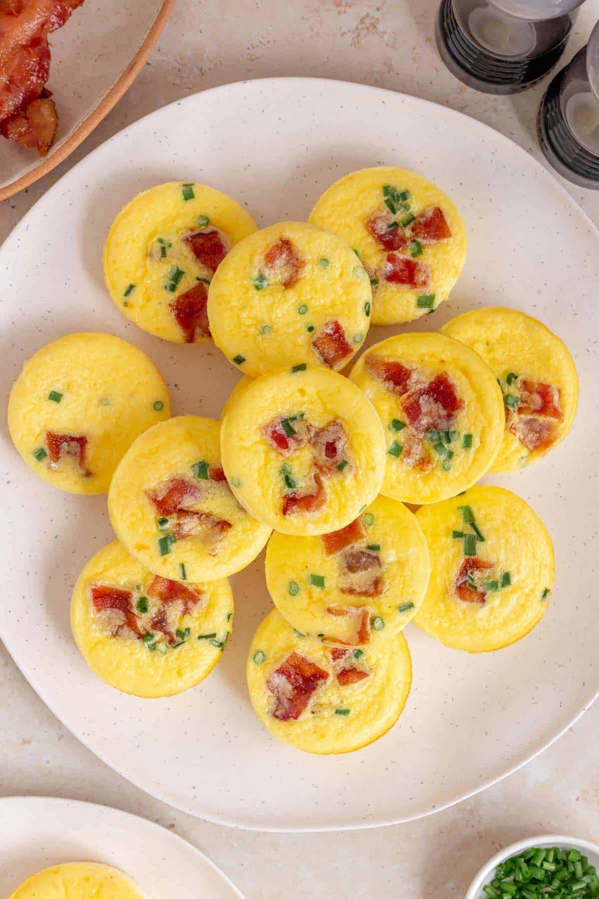 A plate of yellow egg bites garnished with small pieces of bacon and green herbs, arranged in a circular pattern on a white plate. The table surface is light-colored, with a glimpse of other food items nearby.