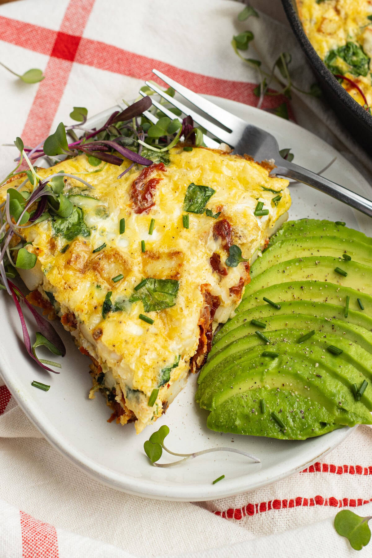 A slice of spinach and sun-dried tomato frittata is served on a white plate with sliced avocado on the side. The plate is garnished with microgreens and a fork is placed beside the frittata. A red checkered napkin is underneath the plate.