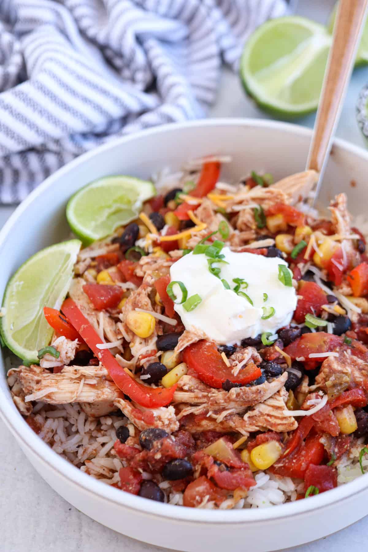 A bowl of shredded chicken, black beans, corn, red bell pepper, and diced tomatoes served over rice. Topped with sour cream and chopped green onions. Lime wedges are on the side. A fork rests in the bowl, and a striped cloth is in the background.