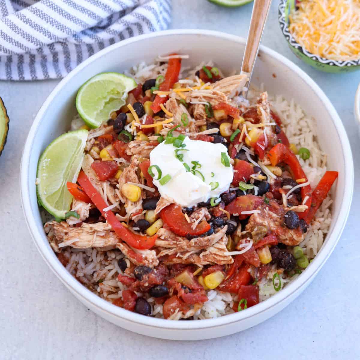 A white bowl contains a colorful Mexican-inspired rice dish. It features shredded chicken, red bell peppers, corn, black beans, diced tomatoes, cheese, sour cream, and lime wedges. A fork rests in the bowl, surrounded by avocado and grated cheese.