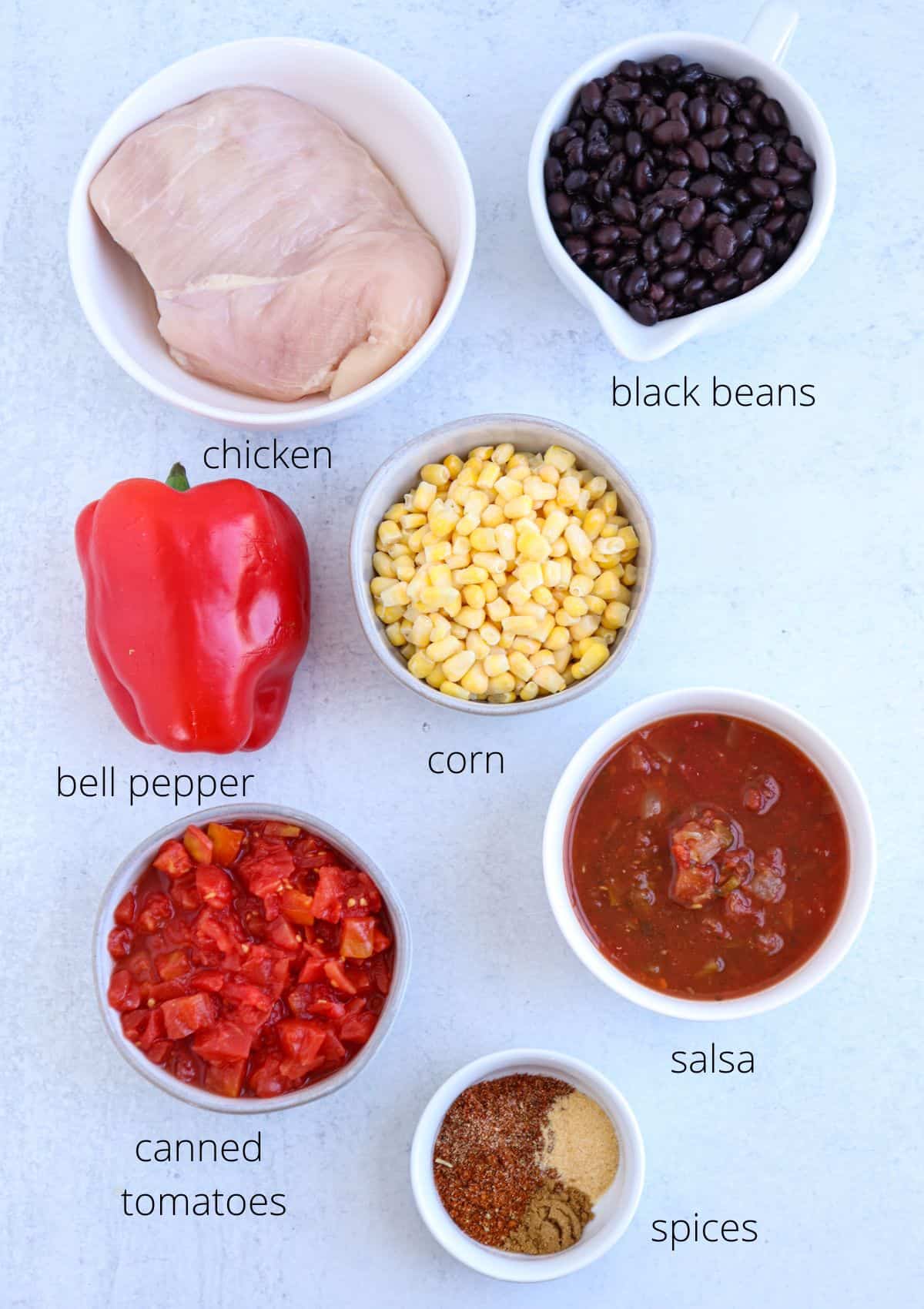 Ingredients for a recipe displayed on a white surface: raw chicken breast, black beans, corn, red bell pepper, canned tomatoes, salsa, and assorted spices each in separate bowls.