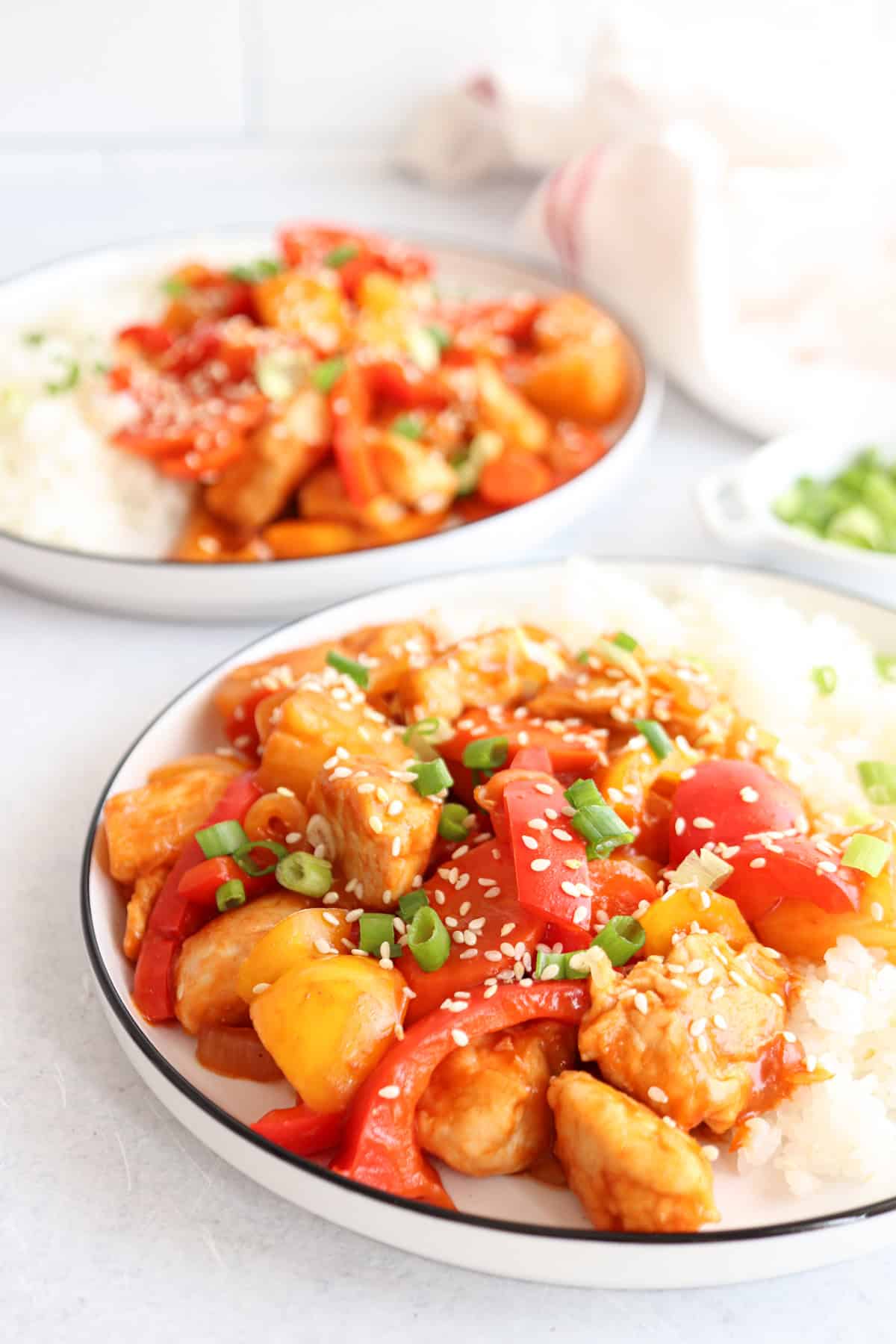 Two plates of sweet and sour chicken are served with white rice. The dish includes pieces of chicken, red and yellow bell peppers, and is garnished with sesame seeds and chopped green onions. Another plate is blurred in the background.