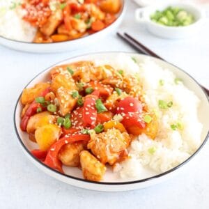A plate of sweet and sour chicken features pieces of chicken, red and yellow bell peppers, and green onions, sprinkled with sesame seeds. Served alongside white rice. Another plate and a small bowl of green onions are in the background.