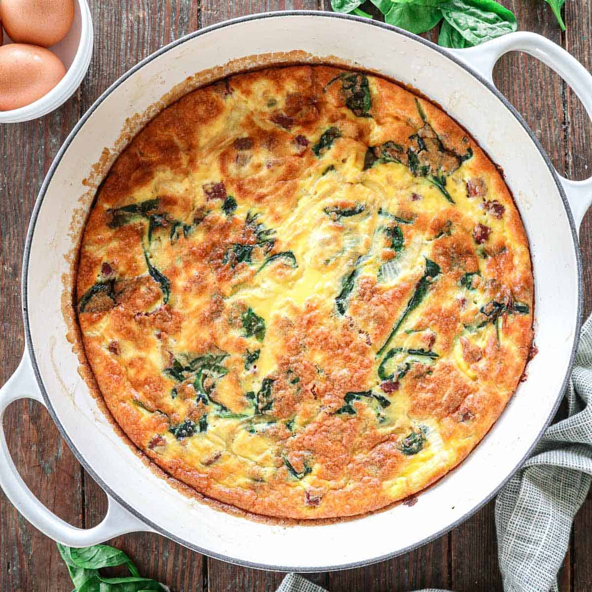 A baked frittata in a round white dish on a wooden table. The frittata is golden brown with visible greens and cheese. Fresh spinach leaves and eggs are nearby, along with a gray cloth in the bottom right corner.