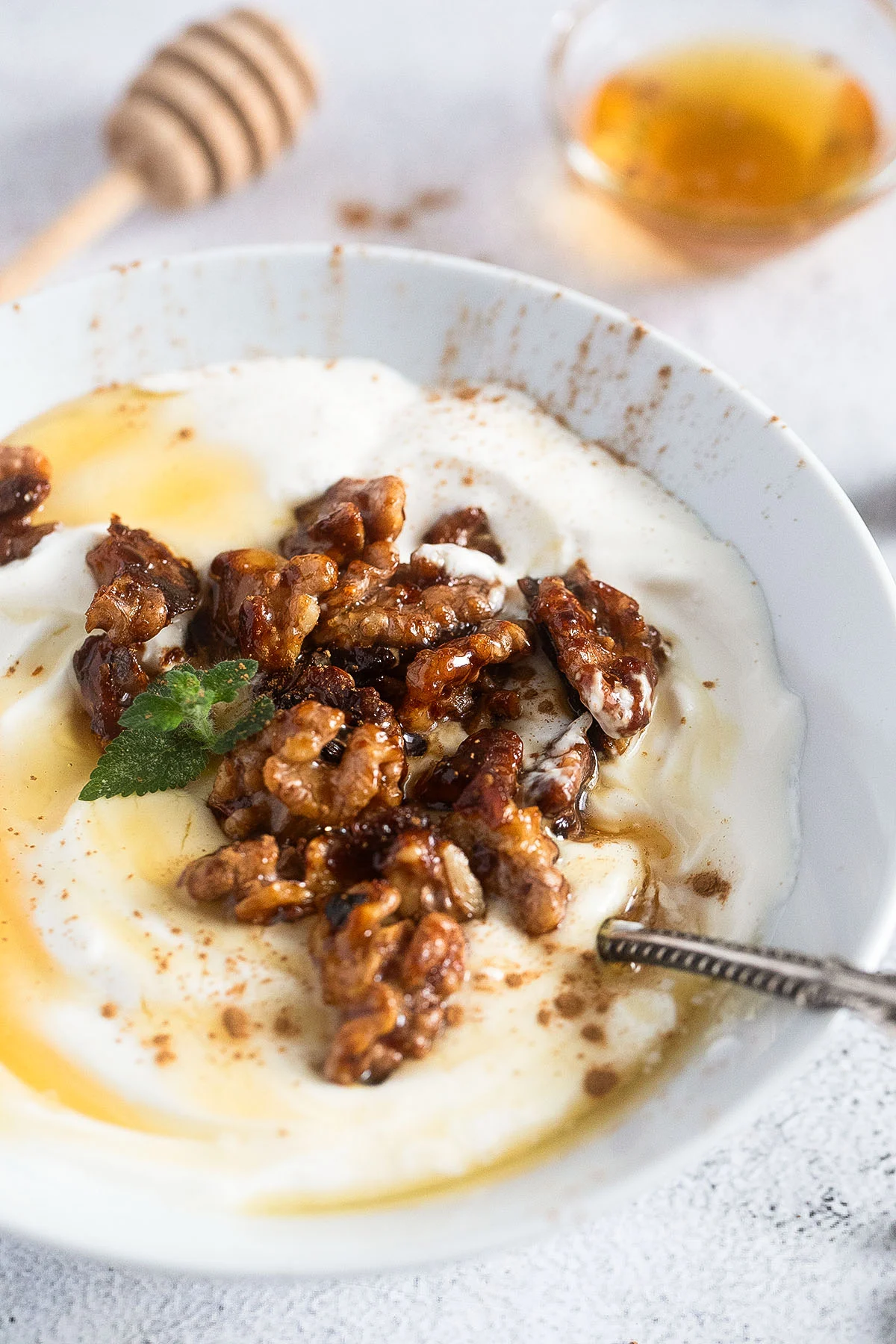 A bowl of yogurt topped with glazed walnuts, a drizzle of honey, and a small mint leaf. A honey dipper and a small dish of honey are in the background. A silver spoon rests in the bowl.