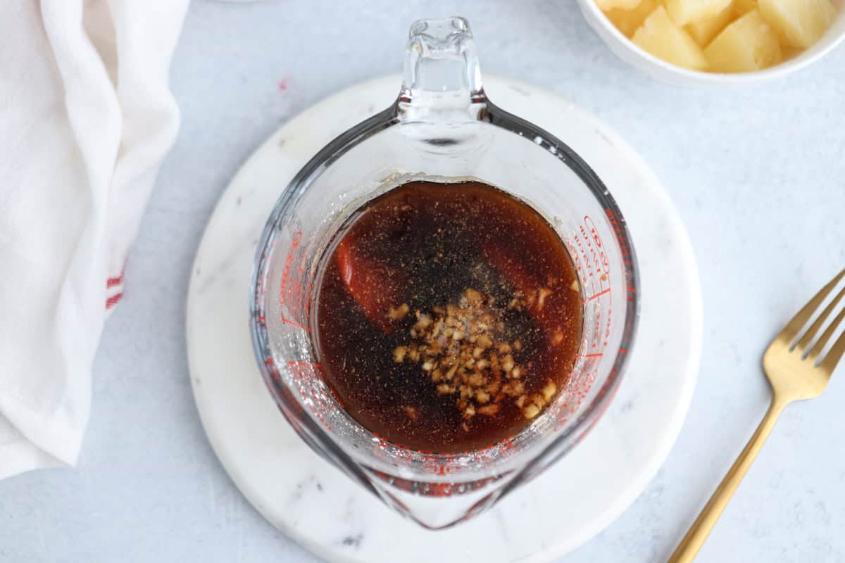 A measuring cup filled with dark sauce and minced ingredients sits on a round white marble board. A fork lies nearby, and a bowl with pineapple chunks is partially visible, along with a white cloth.