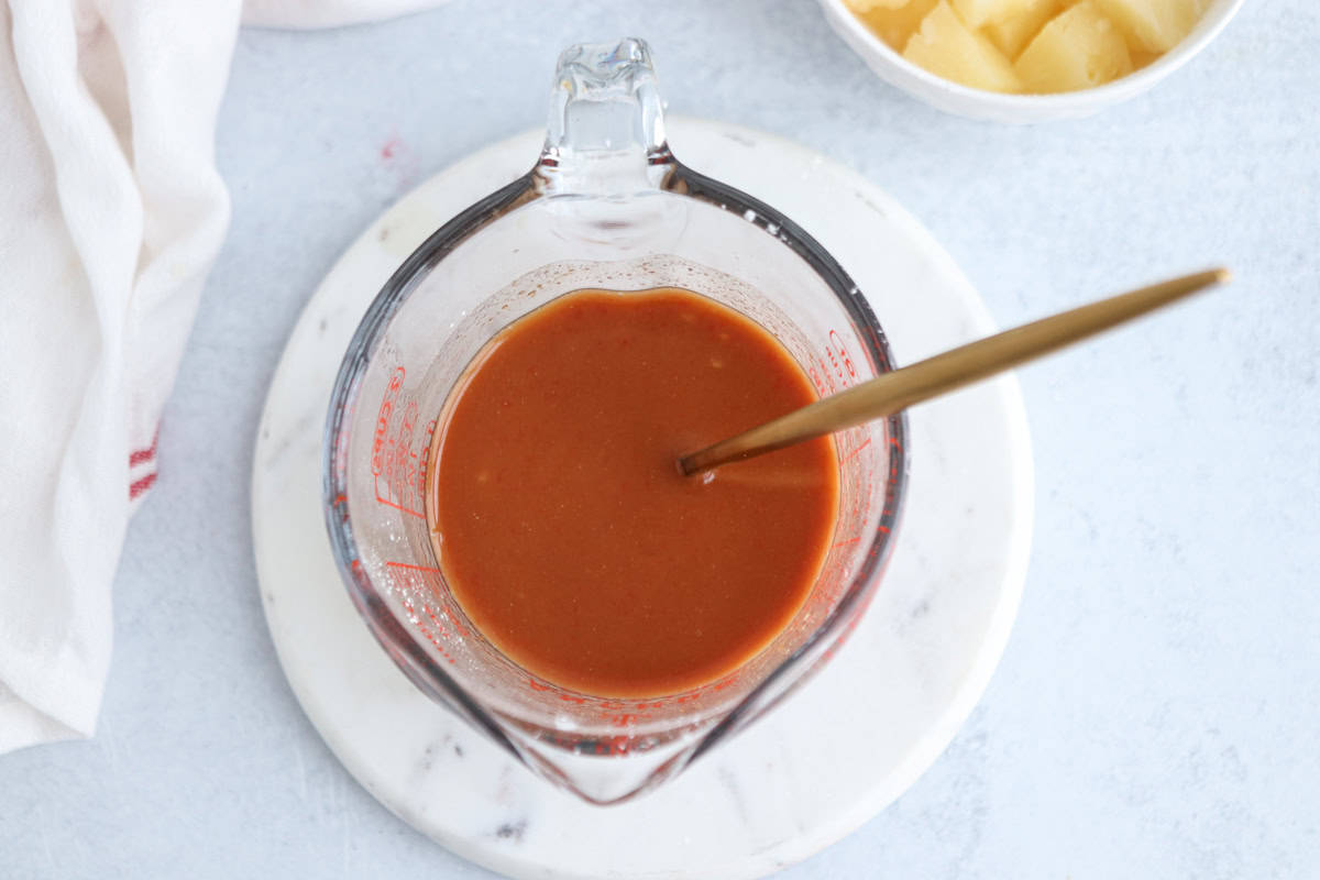 Glass measuring cup filled with a reddish liquid, likely a sauce, and a spoon inside. The cup is placed on a white marble surface. In the background, a bowl with pineapple chunks and a white cloth are visible.