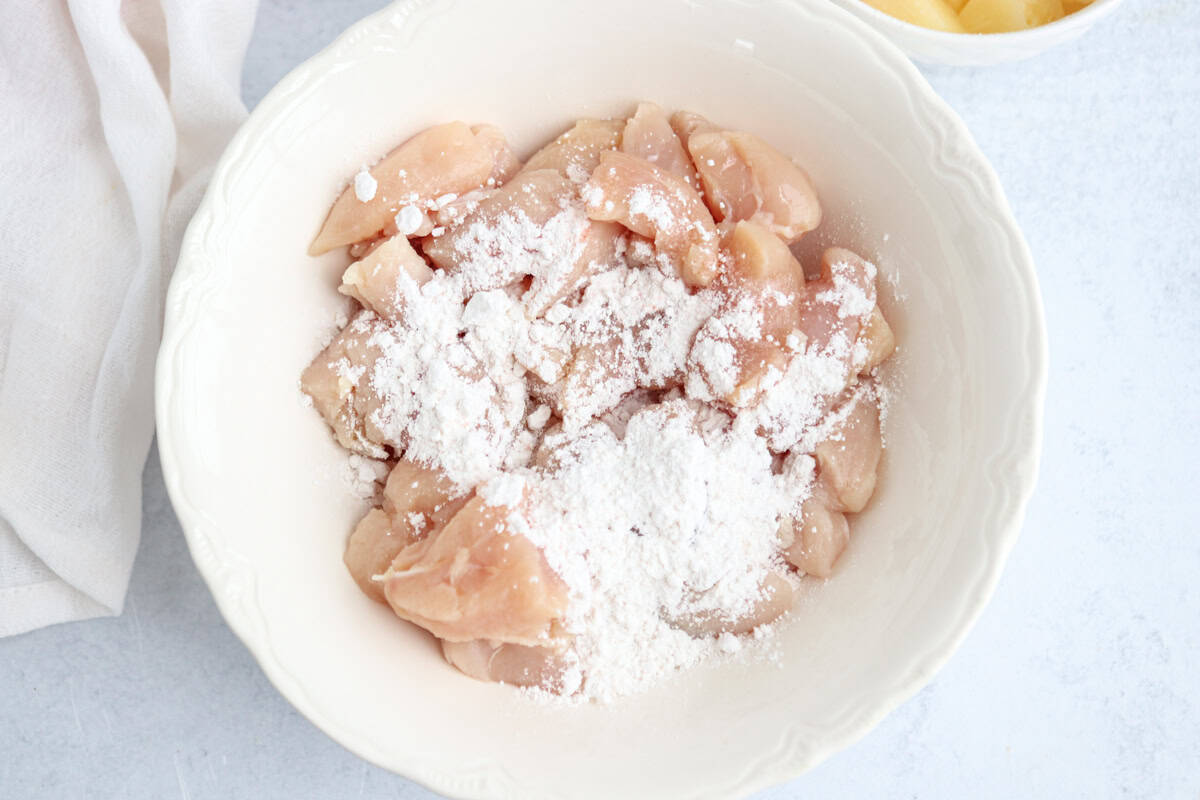 Raw chicken pieces in a white bowl are sprinkled with white flour. A white cloth is partially visible on the left and a small portion of a yellow item is seen in a separate bowl on the right. The background is light gray.