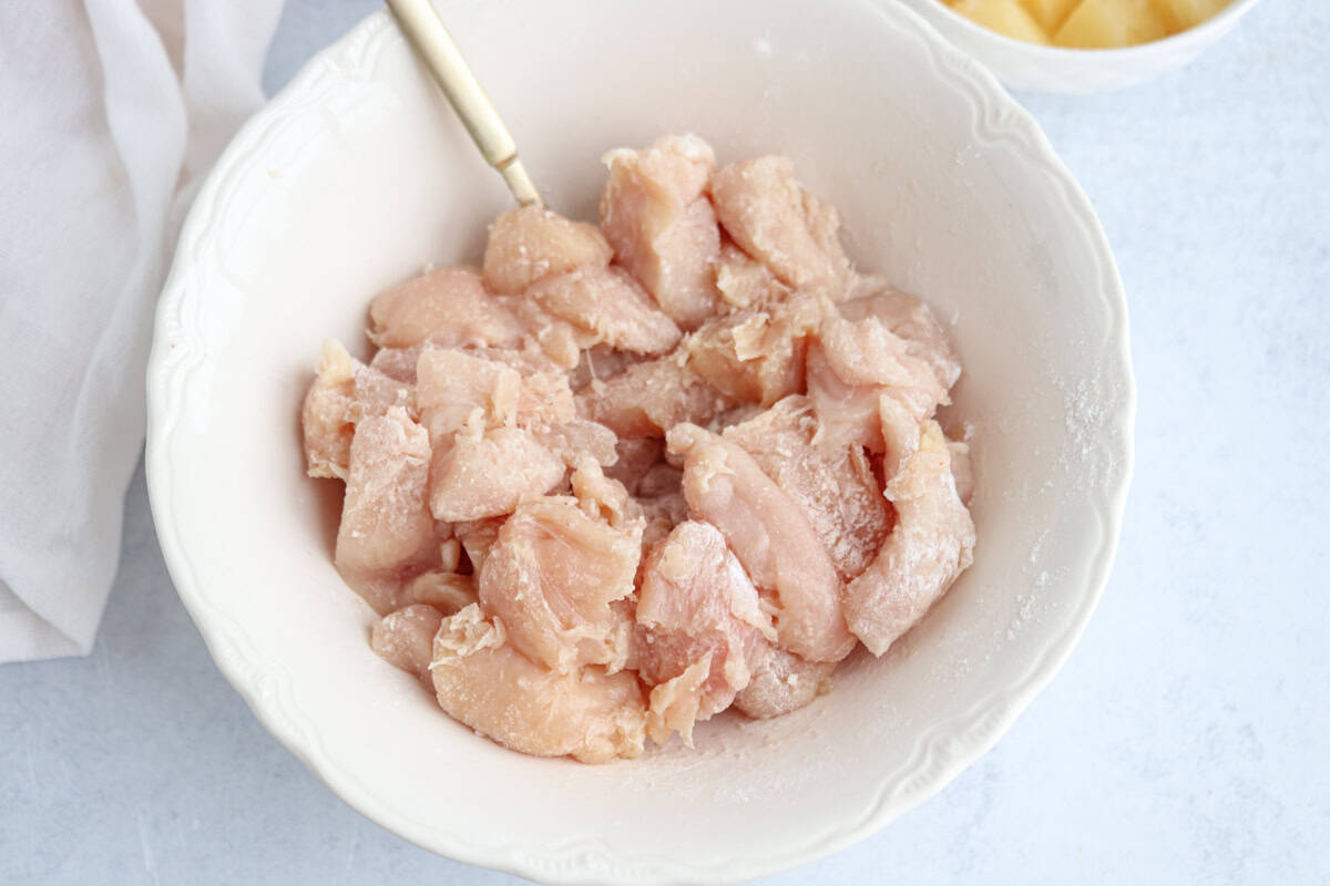 A white bowl contains raw, diced chicken pieces with a spoon, sitting on a light gray surface. A portion of a white cloth and part of a yellow bowl are visible in the background.