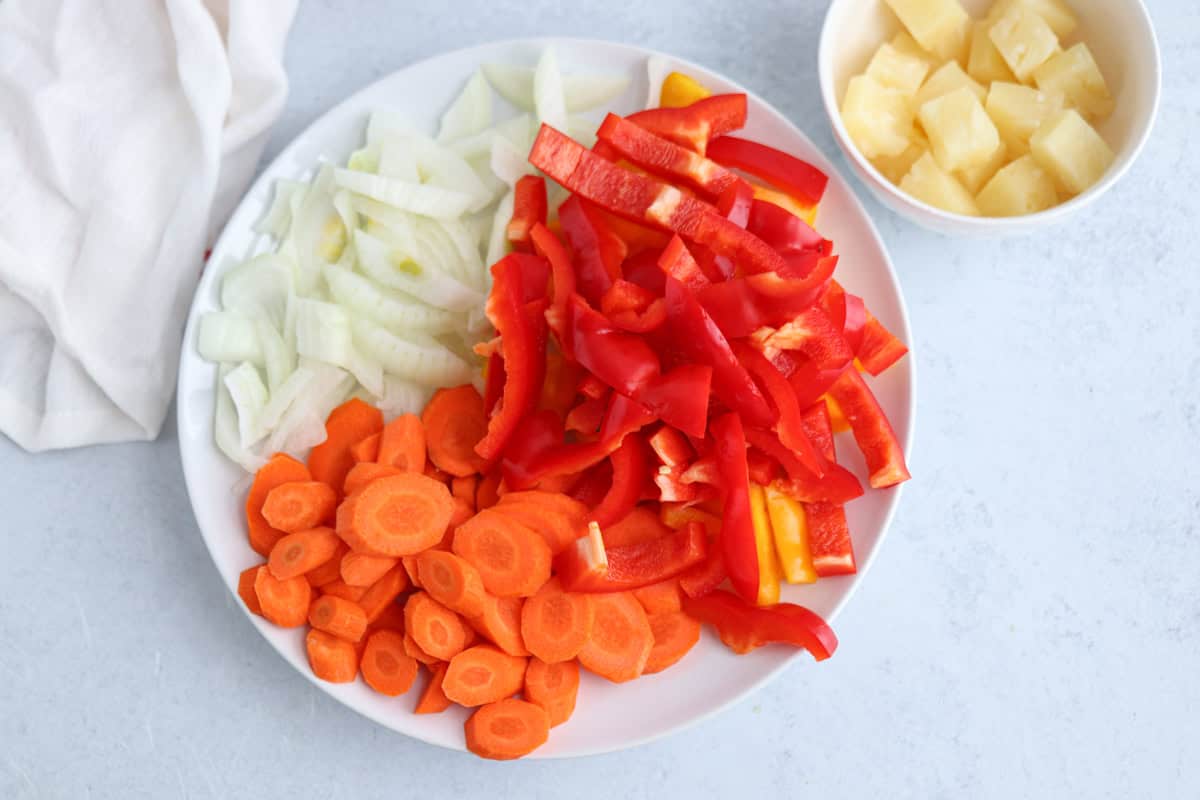 A white plate with sliced onions, carrots, and red bell peppers, placed on a light surface. A small bowl nearby contains pineapple chunks. A white cloth is partially visible on the side.