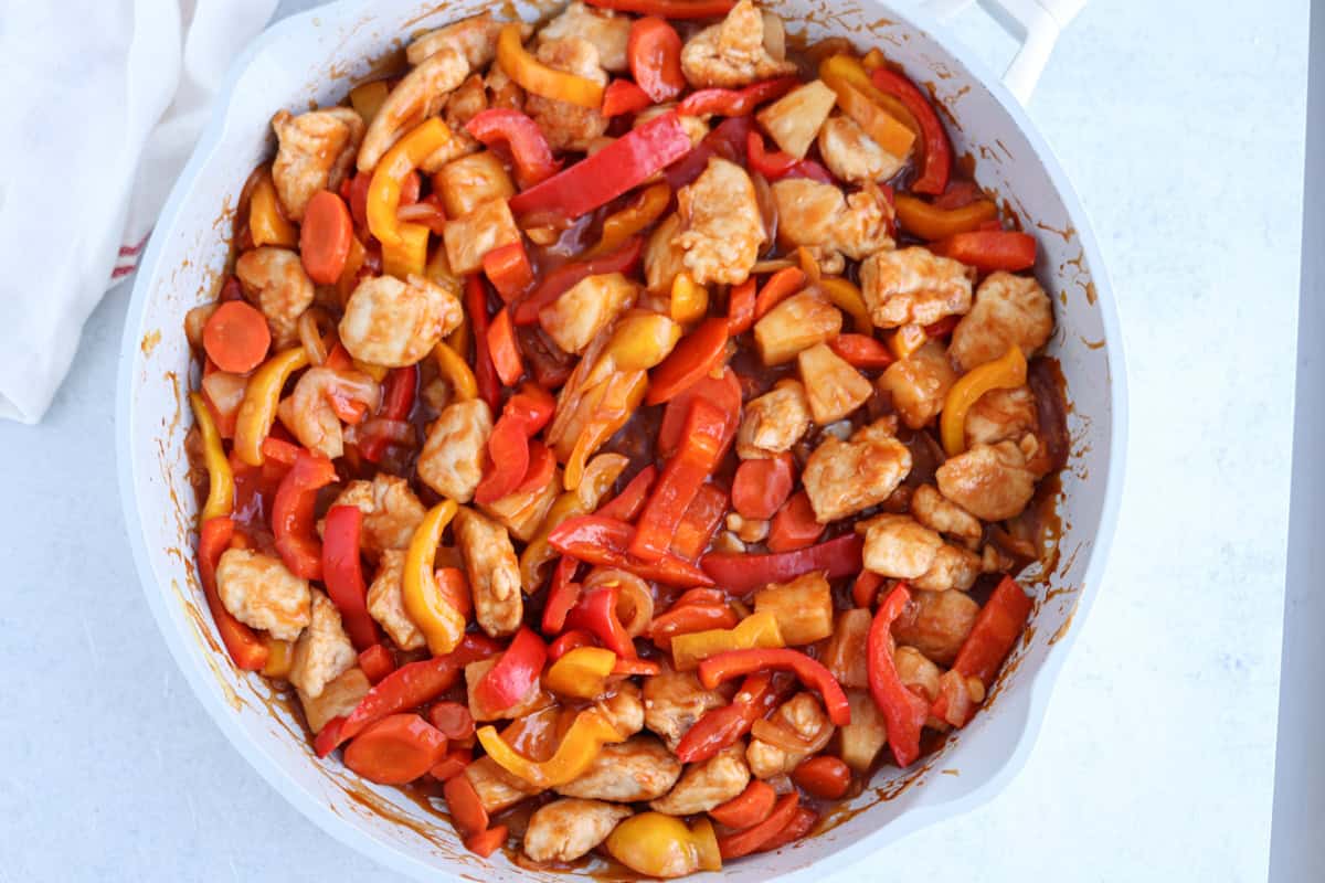 A pan filled with sweet and sour chicken, featuring pieces of chicken, red and yellow bell peppers, and carrots, all coated in a glossy sauce. The mix is colorful, with a blend of orange, red, and yellow hues.