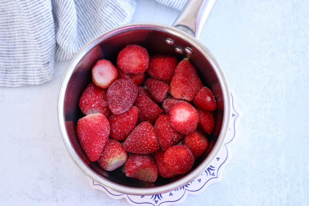 A stainless steel pot filled with frozen strawberries sits on a blue-patterned plate. A striped cloth is adjacent to the pot, placed on a light gray surface.