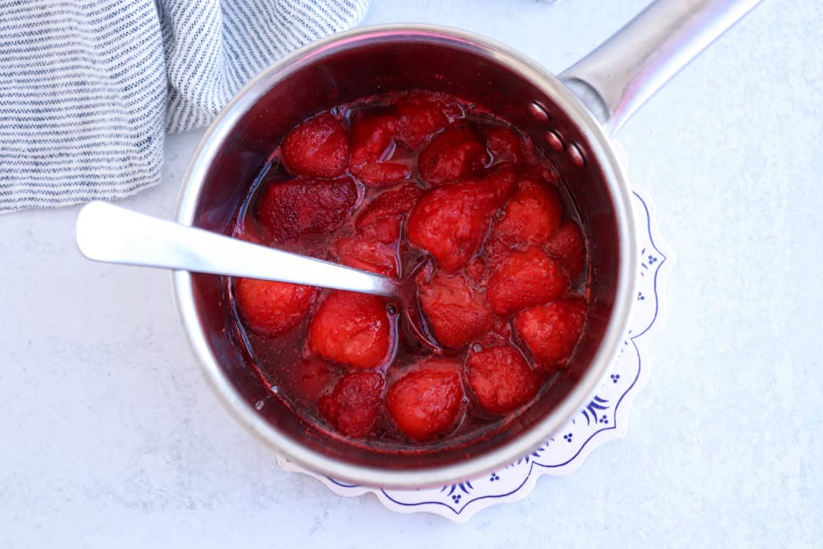 A stainless steel saucepan containing cooked strawberries with sauce. A spoon rests inside the pot. The pot is on a white and blue patterned trivet, and a striped cloth is partially visible in the background.