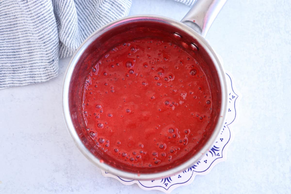 A saucepan filled with bubbling red sauce is placed on a decorative white and blue plate. A striped cloth lies next to the pan on a light gray surface.