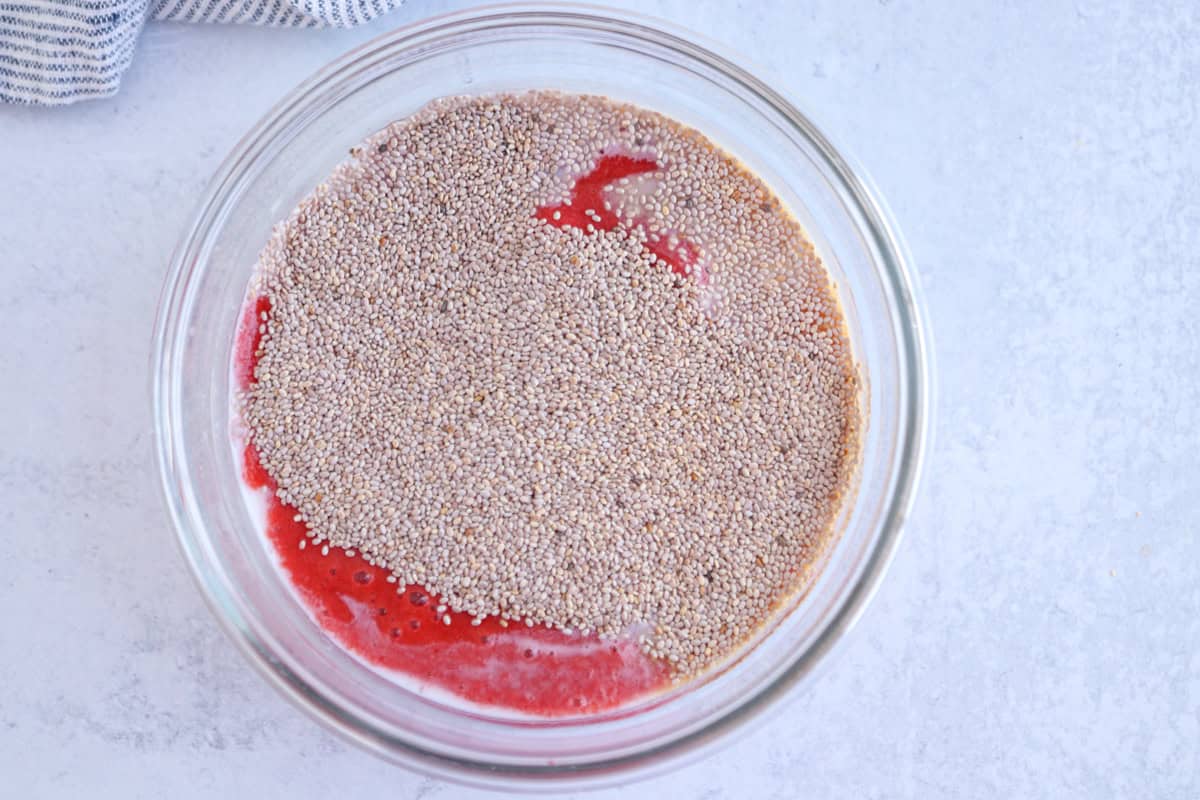 A clear glass bowl contains a mixture of chia seeds soaking in a red liquid, possibly fruit puree, on a light-colored countertop.