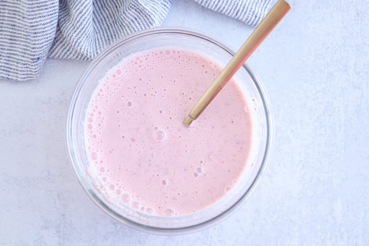 A glass bowl filled with pink smoothie and a gold spoon is on a light-colored surface. A blue and white striped cloth is partially visible in the upper left corner.