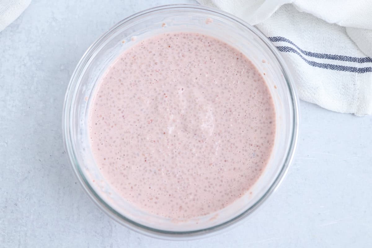 A glass bowl filled with pinkish batter or mixture, possibly containing small air bubbles, sits on a light-colored surface. A white cloth with a blue stripe is partially visible in the background.