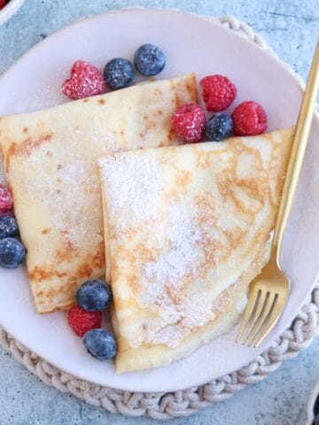 A plate of nalesniki, delicately dusted with powdered sugar, is garnished with raspberries and blueberries. A gold fork rests on the right side of the plate, which sits on a textured mat. The blue surface and bowls of berries in the background complete this inviting scene.
