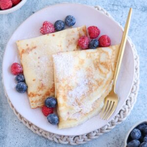 A plate of crepes, reminiscent of a classic nalesniki recipe, is sprinkled with powdered sugar and served with raspberries and blueberries. A gold fork rests on the plate, sitting atop a textured mat, with additional bowls of berries in the background.