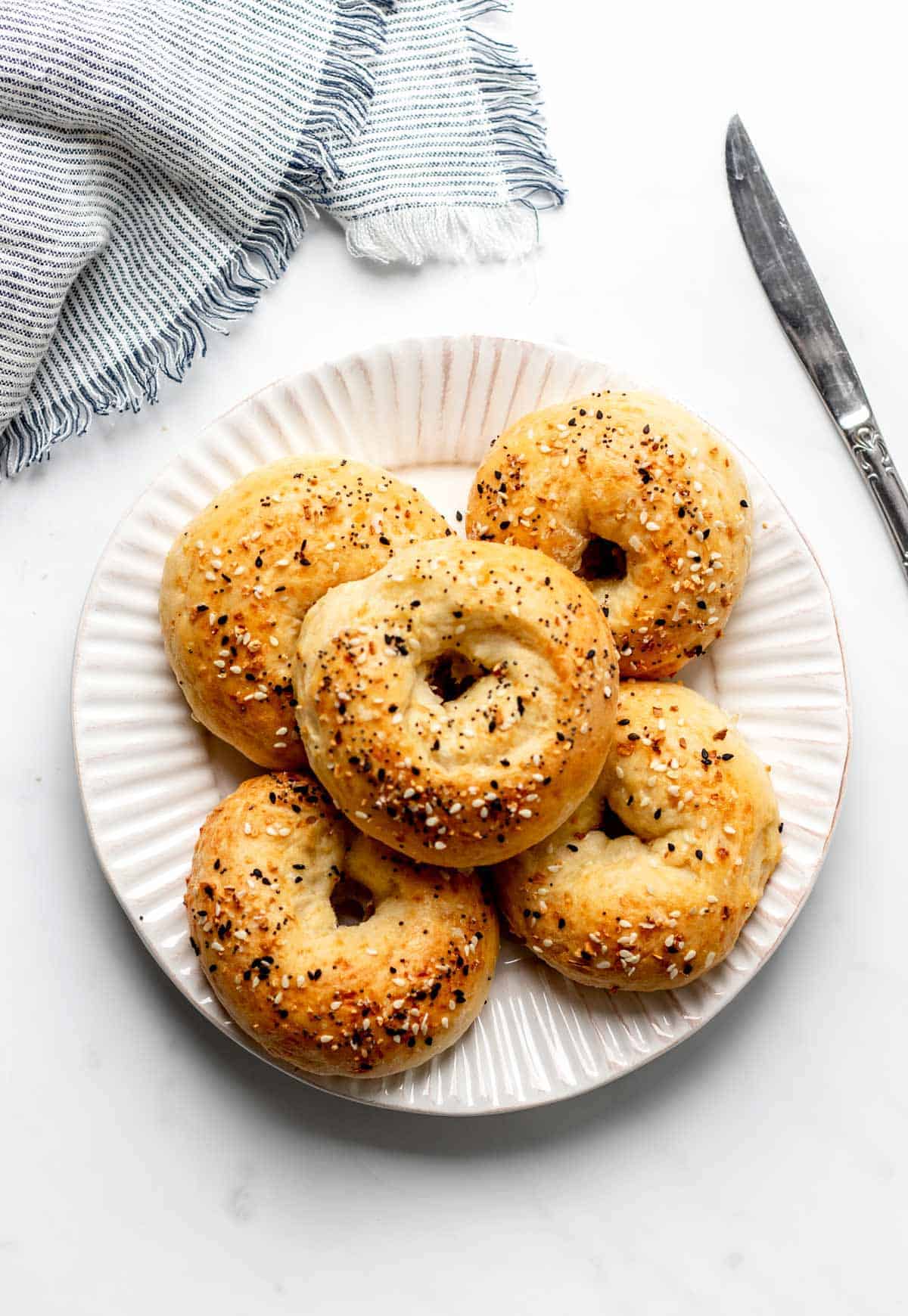 A white plate with five sesame seed bagels arranged on it. A textured blue and white cloth napkin is partially visible next to the plate, and a butter knife is placed alongside. The background is a white surface.