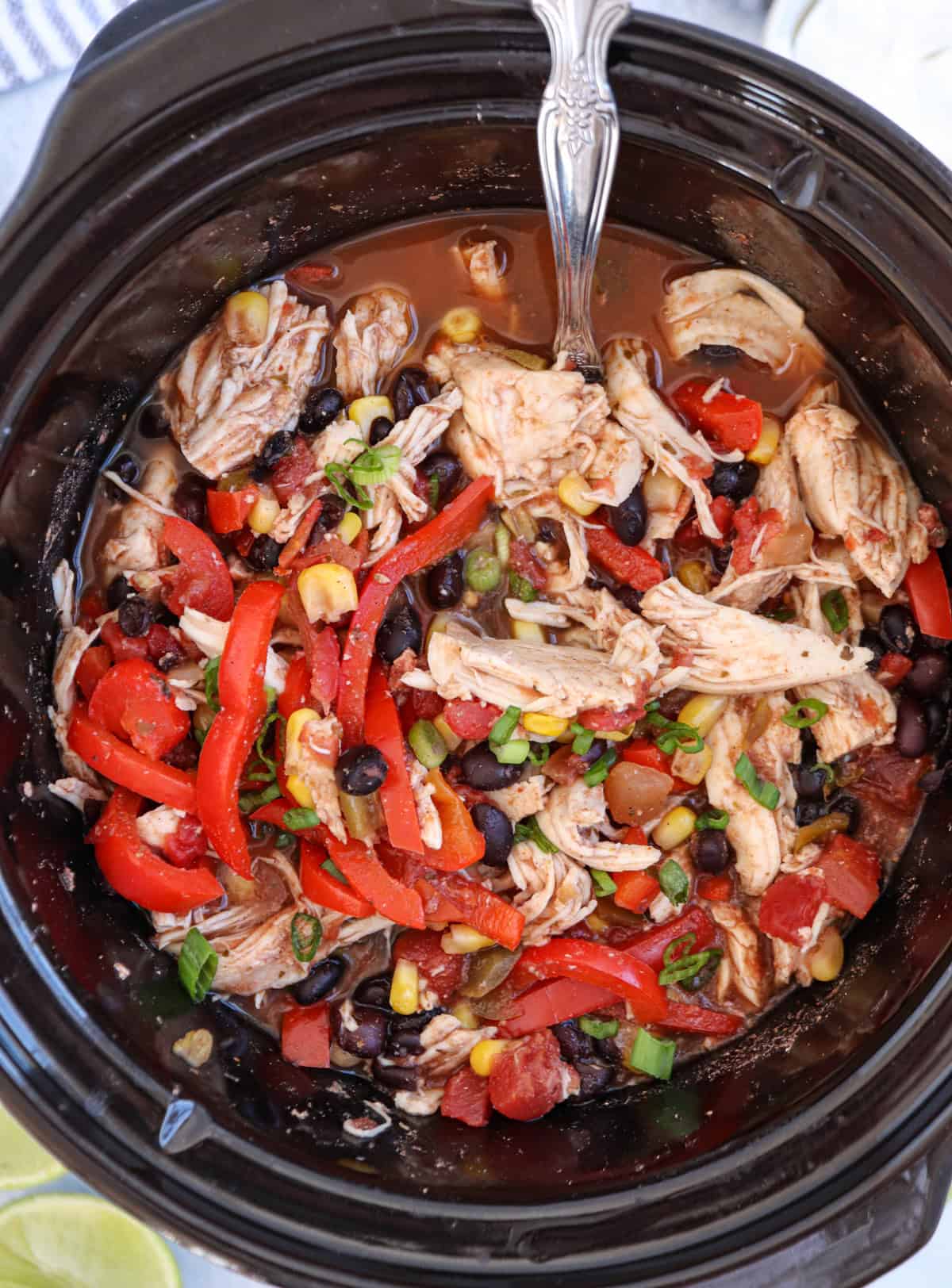 A crockpot brims with a vibrant southwest chicken dish, featuring shredded chicken, black beans, corn, red bell peppers, and chopped green onions. A silver spoon rests within the mix, eager to serve up this delicious meal.